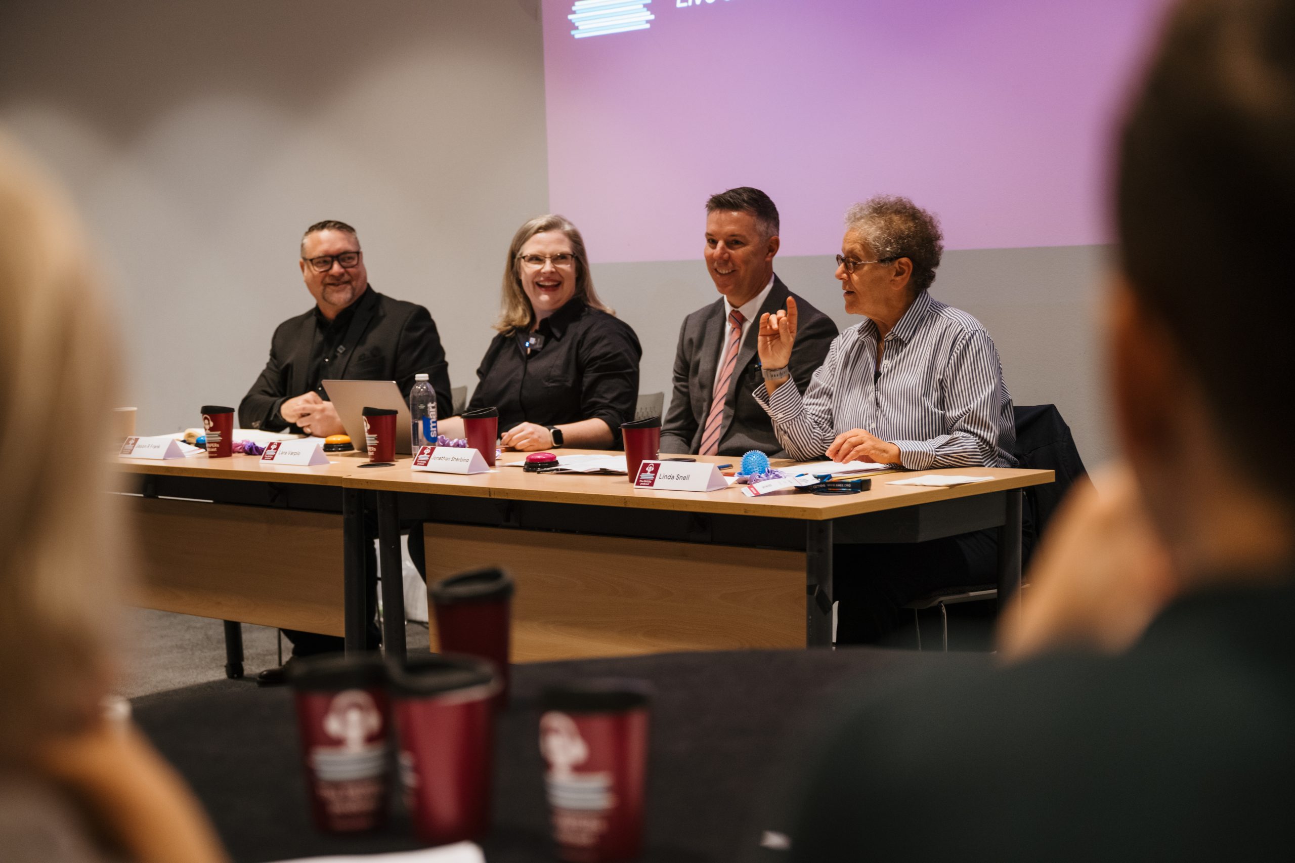 A panel of four engaged professionals sits at a table during a discussion or presentation in front of an attentive audience.