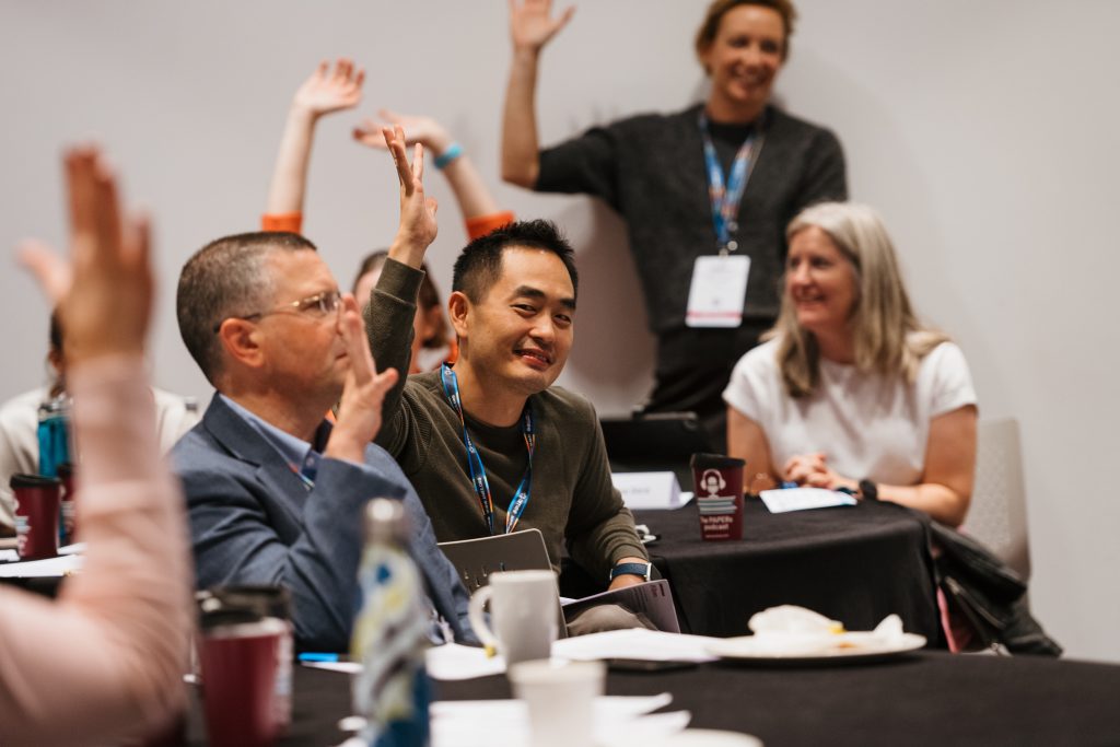 Conference participants interact together by raising their hands, and smiling, one person looks in the camera and smile.