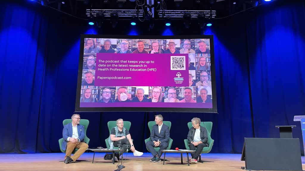 The four hosts during the live session sitting in armchairs on the conference stage with a big screen behind showing Papers podcast marketing material. Headlights are on them.