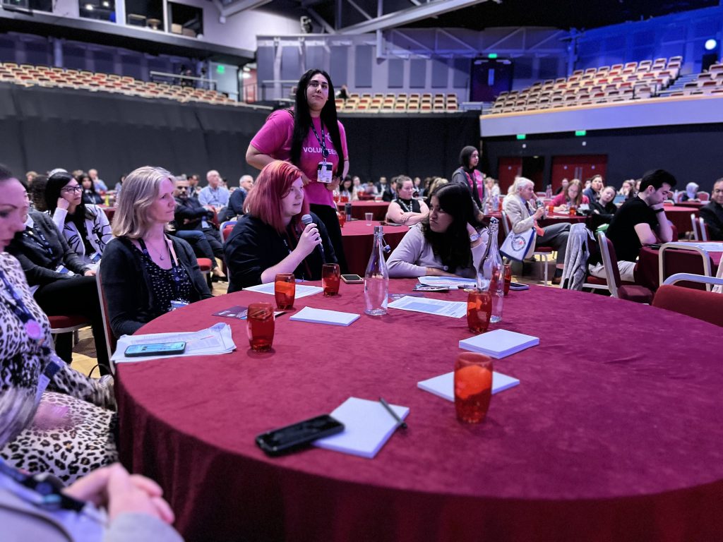 Audience during the live session, sitting at a round table, one person is holding a microphone and asking a question.