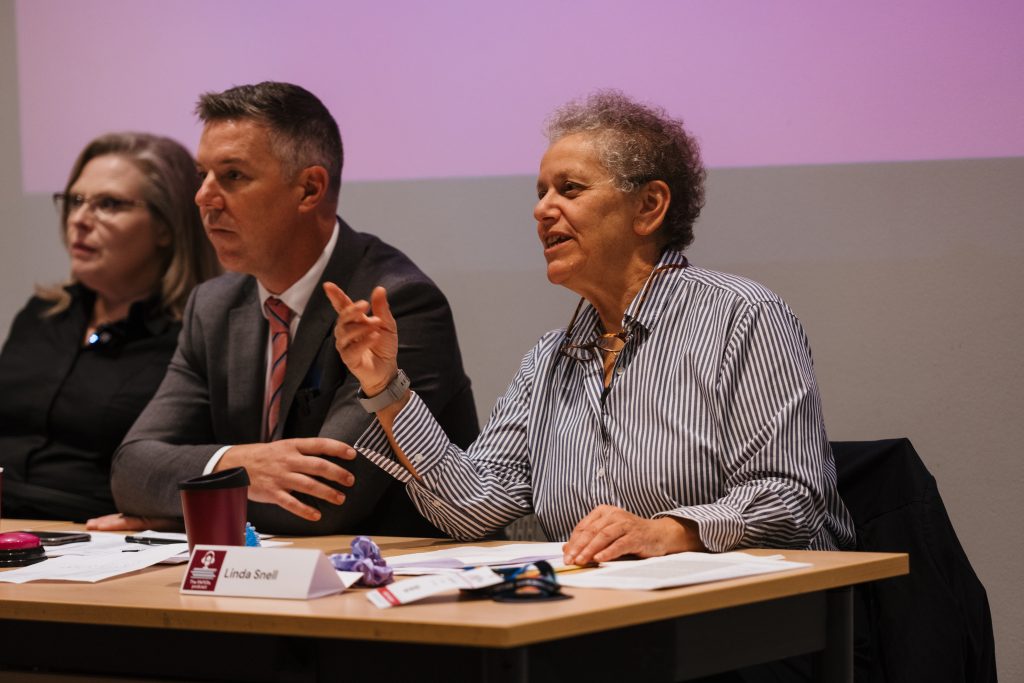 Three persons at a conference table with papers, a coffee mug and nameplates on, looking concentrated, one of them is pointing a finger and saying something.